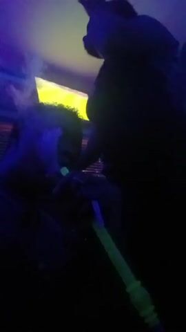 Two people are inside a dark room with colorful lights. One person is smoking from a green hookah. They blow out smoke in the other person's direction. A television screen is visible in the background.