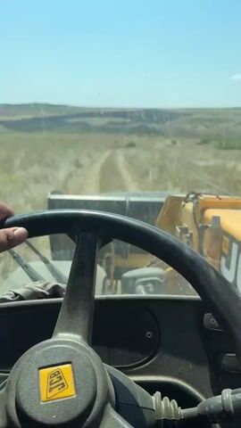 The video shows a first-person perspective from inside a yellow tractor. A person's left hand, wearing a dark watch, is on the steering wheel. The tractor is on a dirt road in a dry, grassy field under a clear blue sky. In the distance, there are hills and a landscape that appears to be a quarry or construction site. The tractor is moving forward.