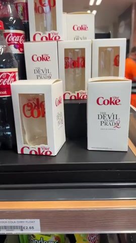 The video shows a close-up of several white boxes containing Diet Coke branded glasses displayed on a black shelf. Each box features the red "Diet Coke" logo at the top and the text "THE DEVIL WEARS PRADA 2 ONLY IN CINEMAS" in black and red lettering below it. Some boxes are open, revealing clear glasses with the "Diet Coke" logo in red. A partially visible price tag below the shelf shows "£2.65". In the blurred background, other products, store fixtures, and people, including one person in an orange high-visibility vest, are visible. The camera slowly pans from left to right across the display.