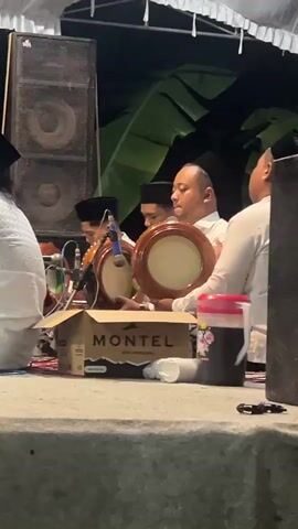 A group of men in white shirts and black caps are playing hand drums. The man in the center holds a drum with a light brown rim and a lighter colored head, and appears to be singing or chanting. To his left, another man plays a similar drum. To his right, another man is visible, also wearing a white shirt and black cap. In the foreground, a cardboard box with the word "MONTEL" is visible. To the right of the box, there is a large dark container with a red lid, and some white paper cups. Behind the men, large speakers are mounted. Large green leaves are visible in the background, suggesting an outdoor setting or a decorated indoor space. The scene appears to be illuminated by artificial light.