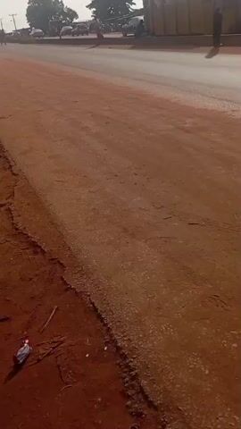A white car is parked on the left side of the road. The road appears to be unpaved with red dirt on the left shoulder and a paved surface on the right. Several people are visible in the distance on the left side of the road, as well as a few vehicles. A large yellow container is situated on the right side of the road. Another white car drives down the paved road from right to left. The sky is overcast and the lighting suggests it is daytime.