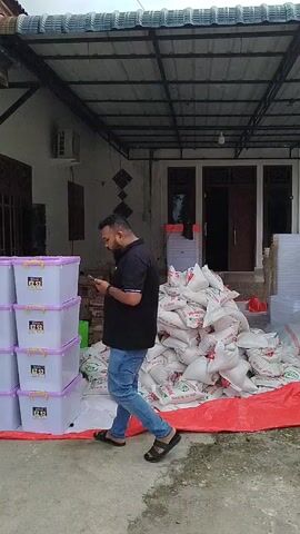 A man in a black polo shirt and blue jeans stands facing away from the camera, looking at a pile of white sacks on a red tarp. To his left, a stack of white and purple boxes are visible. Behind the man, a building with an overhang roof is seen. The man appears to be holding a phone to his ear. The scene is outdoors, with a concrete ground.