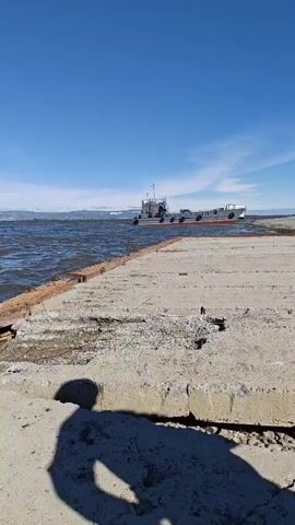 The video shows a concrete pier extending into the water under a clear blue sky. On the water, two barges are visible, one closer to the pier and another further away. On the shore behind the pier, there are several trucks, including a white truck with a trailer full of containers, and a smaller white delivery truck. A tall metal tower, possibly for cranes, is also visible in the background. The sun casts shadows on the concrete pier, indicating it is a sunny day. The water appears choppy, suggesting some wind.