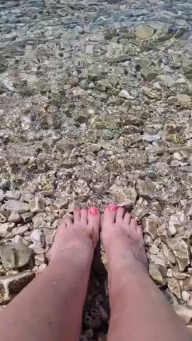A POV shot shows a pair of bare feet with bright pink toenail polish submerged in clear, shallow water. The water is moving, creating ripples that distort the view of the rocky seabed below. The rocks are varied in size and color, predominantly shades of grey and white. A hand with matching pink nail polish is visible reaching towards the feet, touching one of the ankles. The scene appears to be a beach or a shallow riverbed on a sunny day.