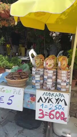 This video shows an outdoor market stall with various items for sale, including pies and drinks. A person in a black shirt and shorts is standing behind the counter, possibly serving customers. There are potted plants and flowers around the stall, adding to the vibrant atmosphere. In the background, a white car is parked on a cobblestone street, and a bell tower is visible in the distance. The sky is clear and blue, indicating a sunny day.