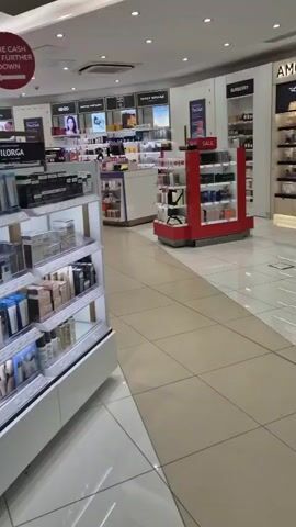 The video shows a view of a beauty store. In the foreground, there are shelves stocked with various beauty products, including makeup and skincare items. Brands like Dermalogica and Filorga are visible on signage. Further in the background, more aisles of products are visible, along with signage for brands like Clarins, Kérastase, and Mugler. A red sign with white text saying "MORE CASH TILLS FURTHER DOWN" is hanging from the ceiling. The store appears well-lit and organized.
