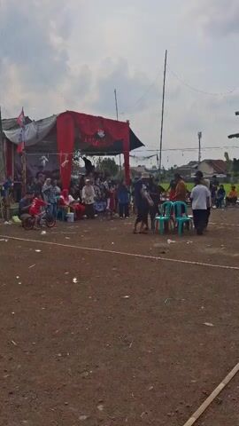 A group of people are gathered outdoors under a cloudy sky. A red tent is set up in the background, adorned with banners and draped fabric. A crowd of spectators is visible behind a barrier, some seated and others standing. In the foreground, several individuals are interacting, with some seated on plastic chairs around a table. Two bicycles are parked to the left of the scene. The ground is covered in dirt with scattered debris. The overall impression is that of a local gathering or event taking place in an open area.