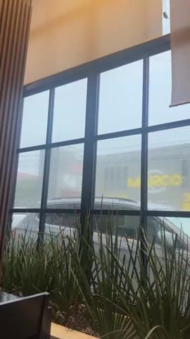 A slow camera pan shows a view through a large window onto a street in what appears to be a restaurant or cafe. It is raining, with water droplets visible on the windowpane and on the roof of a parked car. Outside, a silver SUV is parked on the street, and a yellow sign with the word "MARCO" is visible on a building across the street. In the foreground, a planter filled with green, spiky plants separates the window from the interior of the establishment. To the left of the window, a wall paneled with vertical wooden slats is visible, along with dark chairs and a table. The camera pans to the right, revealing more of the interior seating area, including a row of dark chairs against a wooden paneled wall.