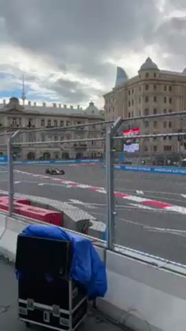 The video shows a Formula One race car driving along a race track in an urban environment. The race car is red and blue and the track has red and white lines. There is a tall building with a dome in the background, as well as other buildings and structures. A chain-link fence with "AMERICAN EXPRESS" on the side is in the foreground. In front of the fence is a road case covered with a blue tarp. The sky is overcast.