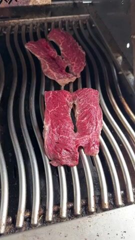 Two pieces of raw meat, likely steak, sit on the metal grill of a barbecue. The lower piece is shaped like a rectangle split down the middle, while the upper piece is smaller and oddly shaped. The grill grates are clean and shiny. The meat is bright red, indicating freshness. The grill itself appears to be stainless steel.