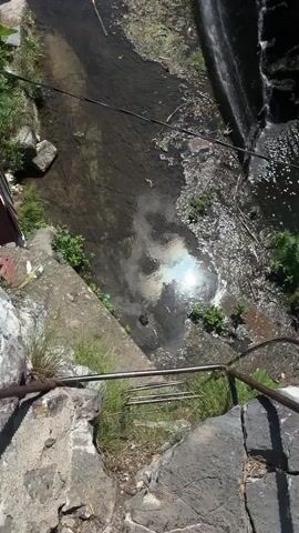 The video shows a view from above of a waterfall and a body of water. In the upper left portion of the screen, three people can be seen standing in the shallow water near some stone steps. The water itself appears somewhat murky with debris floating on the surface, including what looks like algae and branches. A concrete structure, possibly a path or walkway, is visible along the left bank, with a small brown building with a red roof peeking out from the corner. Sunlight glints off the surface of the water, creating a shimmering effect. To the right, a waterfall cascades down a rocky cliff face into the water below. The surrounding vegetation is lush and green, with trees and bushes growing along the banks and on the rocks. The scene suggests a natural outdoor setting, possibly a park or a recreational area, on a sunny day. The presence of people suggests it is a warm time of year. There is no visible text in the video.