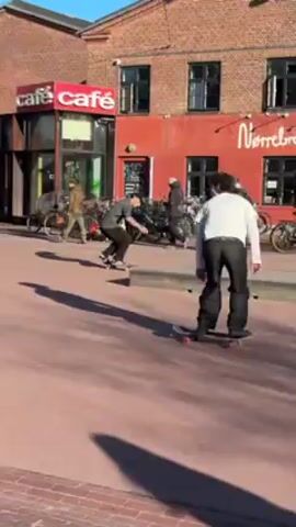 A skateboarder wearing a white shirt and black pants approaches a low concrete platform on a red brick plaza and successfully performs a skateboarding trick on the platform. A cafe is visible in the background. Other people are visible in the background, and a dog is standing nearby. The sky is blue.