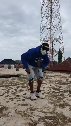 A person wearing a blue hoodie, denim shorts, and goggles stands on a rooftop in front of a tall communication tower. The person performs a brief dance routine.