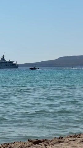The video captures a bright, sunny day at a beach. The foreground shows a sandy shore with some pebbles and a few small rocks. The middle ground is dominated by a clear, turquoise sea with gentle waves rippling towards the shore. In the distance, a large white yacht is visible on the left side of the frame. To the right of the yacht, a person is water skiing behind a small boat, creating a spray of water. Further to the right, another person on a watercraft is also visible. In the background, a hazy, green-covered hill or mountain range forms the horizon under a clear, pale blue sky. The overall scene depicts a leisurely summer day with water activities.