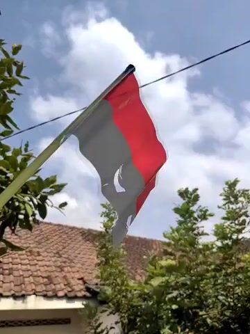 A flag with red and gray stripes and a hole in the middle is blowing in the wind. The flag is attached to a pole. The background shows a blue sky with clouds, trees, and the roof of a building.