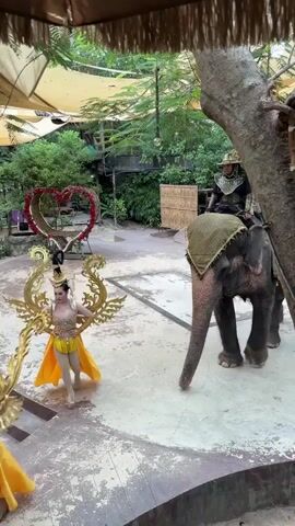 A woman in a golden, elaborate costume with feathered headdress and a yellow skirt stands on a platform with her hands extended to her sides. To her right, an elephant walks forward, carrying two people. The person in front of the elephant wears a dark, ornate costume and headwear. Behind them, a woman in a gold and yellow outfit sits on a decorated saddle on the elephant's back. She is wearing high-heeled shoes and a headpiece. The elephant is decorated with a patterned cloth on its back and head. In the background, there is a large heart-shaped decoration adorned with red flowers. The environment appears to be outdoors, with trees and foliage visible, and a thatched umbrella-like structure is overhead. The ground is a textured, light-colored surface with some darker patches. The scene is brightly lit.