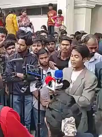 A young man in a gray blazer is surrounded by a crowd of people and is being interviewed by reporters holding microphones.  The crowd includes men of various ages, some holding phones to record the scene.  Several reporters are holding recording equipment and microphones near the man. The setting appears to be outdoors near a building.