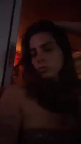 A woman with long dark hair is looking directly at the camera. The lighting is dim, with an orange hue. She is wearing a strapless top. The background is blurry, but there appears to be a door frame and some decorative items. She runs her hand through her hair.