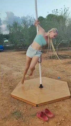 A blonde woman with braided hair is performing a pole dance routine outdoors on a portable stage. She is wearing a light blue crop top and denim shorts. The stage is hexagonal and made of wood. Two red sandals are lying on the ground in front of the stage. The background consists of dirt, gravel, some sparse trees and vegetation, and a few vehicles in the distance. The sky is visible, and it appears to be late afternoon or early evening. The woman is mid-movement, twisting and bending around the pole.