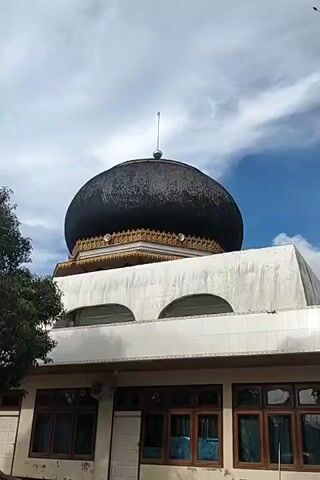 The video shows the exterior of a building, presumably a mosque, featuring a prominent black dome. The dome is accented with gold trim at its base and topped with a thin spire. The building itself has a white facade with arch-shaped windows on the upper level and multiple standard windows on the ground floor. A large green tree obscures the left side of the building and a black car is parked in front of it. The sky above is partly cloudy.