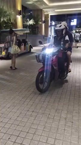 A pink scooter with two people on it, a driver wearing a white helmet and black and green shirt, and a passenger wearing a white cap, rides past a building with "JUNGCEYLON TOURIST SERVICE CENTER" written on it. Several cars are parked in front of the building. The ground is paved with interlocking bricks. The scene appears to be at night, with artificial lighting illuminating the area.