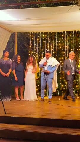 The video shows a wedding ceremony taking place outdoors under a canopy adorned with white fabric and fairy lights. On a raised wooden platform, a man in a blue suit and a white tallit (Jewish prayer shawl) holds a microphone and speaks. Beside him, a woman in a white wedding dress stands with her hand to her face, appearing emotional. To their left are two women, one in a dark blue dress and the other in a shimmering dark purple dress and head covering. To the right of the groom is a man in a grey suit, clapping. The background features greenery and more fairy lights. The video captures the emotional climax of a speech, likely the groom's vows or a blessing, as the bride reacts with tears.