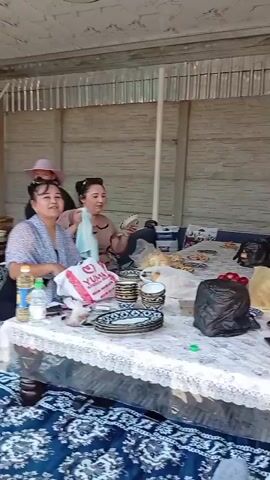 A group of women are gathered around a table spread with food and plates. Some of them are dancing and gesturing with their hands. The setting appears to be an outdoor covered area, possibly a patio or veranda, with corrugated metal walls and a patterned ceiling. One woman in the foreground is wearing a white tank top and black pants, while another in the background is in a blue outfit. There are plastic bags, plates, cups, and food items on the table. The mood seems festive and celebratory.