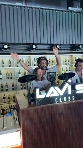 A man with dreadlocks and a woman with blonde hair are seen in the foreground, with the man raising his arms in the air. They are both smiling. Behind them, a man with headphones is visible. The background features shelves filled with bottles, possibly wine. There is a DJ setup with a sign that reads "LAVI CLUBS". The lighting is dim, and there are some lights mounted above the shelves. The overall scene appears to be a performance or event at a club or bar.