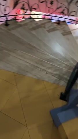 The video shows a spiral staircase with marble steps and a wrought iron railing. The railing has intricate floral designs. Colorful lights are visible behind the railing, casting a pink and blue glow on the stairs. The camera is moving downwards, giving a first-person perspective of descending the staircase. The floor at the bottom of the stairs appears to be tiled in a yellowish hue. A small section of a blue garment is visible in the lower right corner of the frame. The walls appear to be a neutral color, and there is a large mirror or window reflecting the environment.