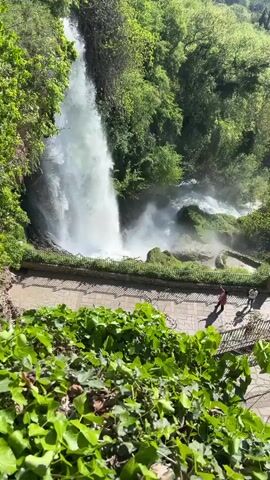A wide, long shot captures a waterfall cascading amidst lush green trees, with a stone pathway visible below the waterfall. A person wearing red stands on the pathway. The camera tilts up to reveal a clear, blue sky and an expansive landscape of green fields and trees extending into the distance. The scene is bright and sunny, creating a clear view of the waterfall and surrounding environment.