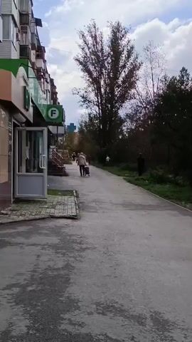 The video shows a pedestrian walkway next to a building with shops. In the foreground, a woman in dark clothing and a jacket is walking away from the camera, carrying two large patterned shopping bags. She appears to be touching her head with her left hand. To her left, further down the walkway, another person with a shopping cart is walking towards the camera. The building has several windows, and one of the shops has a sign that reads "ДЕТСКАЯ ОДЕЖДА ОБУВЬ" which translates to "CHILDREN'S CLOTHING SHOES". The walkway is paved, and there is grass and trees on the right side of the path. The sky is cloudy. The camera is panning slightly to the right.