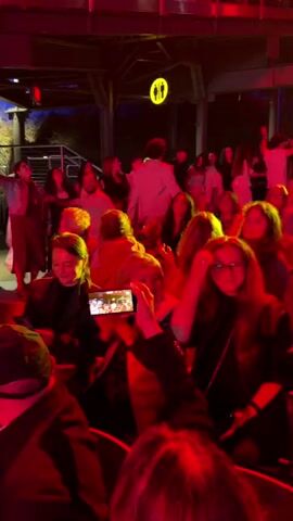 The video shows a crowd of people in an outdoor venue, possibly a concert or performance. The lighting is predominantly red and yellow. In the foreground, a person in a black jacket is filming the event with a smartphone. In the background, people are dancing and waving their arms. Some individuals are holding up flags. There is a sign that reads "NO SMOKING". The overall atmosphere appears festive and energetic.