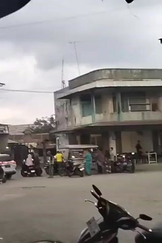 A wide shot of a street scene with several motorcycles and a few people. In the foreground, a black motorcycle is parked. In the middle ground, a group of people are gathered around a food cart. A few motorcycles are parked near the food cart, and one person is riding a motorcycle past. In the background, there is a dilapidated building. The sky is overcast.