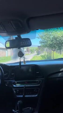 The camera is positioned inside a parked car, with a view of a quiet residential street through the windshield. The street is lined with houses and trees, and a grassy field stretches out in the distance, leading to a gentle mountain range. A couple of pedestrians are walking down the street, heading away from the car. The car's dashboard is visible, featuring a rearview mirror with decorative tassels, a central display screen, and a row of buttons and controls. The sky is bright blue, suggesting a clear and sunny day.