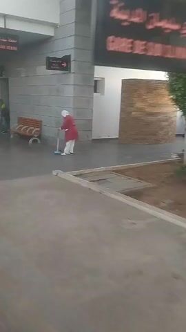 The video shows an outdoor scene, likely in front of a building. There is a sign that reads "GARE DE SIDI SLIMANE MEDINA" in both Arabic and French. A person in a red and white outfit is using a mop near a building. The surrounding area is paved and there are trees and bushes. A sign with a large "H" is also visible. The camera appears to be moving.