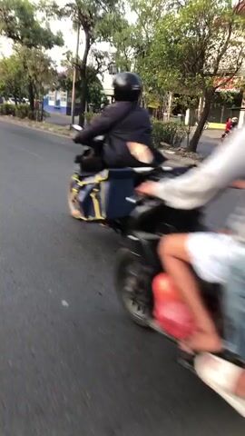 Two individuals, viewed from behind, ride on a motorcycle on a paved road. The rider in front wears a dark shirt and a helmet, while the passenger behind wears a blue shirt and jeans, also with a helmet. The motorcycle appears to be a scooter-style model with a luggage rack at the rear. Another motorcycle is visible ahead, also carrying two people. The environment suggests an outdoor setting with trees lining the roadside and what appears to be a sidewalk or median. The lighting indicates it is daytime, possibly late afternoon due to the warm tones. The road surface is asphalt, and the motion blur suggests the video is captured while moving.