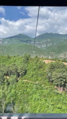 The video shows a view from a cable car ascending a mountain. The cable car is moving forward and upward, as indicated by the changing perspective of the scenery. In the foreground, there are lush green trees and vegetation covering the mountainside. The path of the cable car is marked by two parallel cables extending upwards into the frame. In the mid-ground, a tall pylon supporting the cables is visible. In the background, large, imposing mountains with rocky peaks rise against a bright blue sky with scattered white clouds. The overall impression is of a scenic journey through a mountainous, forested region on a clear day. There are no people or specific actions other than the movement of the cable car itself.