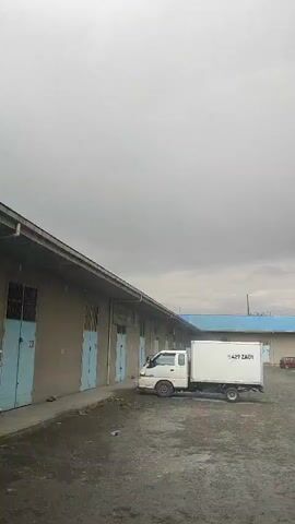 The video shows a warehouse with a white truck parked outside. The truck has a cargo box with writing on the side. The warehouse has several doors that are painted blue. The sky is cloudy and the ground is wet, suggesting that it may have recently rained. The video is taken from a low angle, looking up at the warehouse and the sky. There is no movement in the video, suggesting it is a still image or a very short clip.