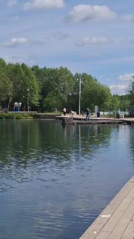 The image displays a serene outdoor scene at a lake or large pond in Kipen, Russia, under a bright, partly cloudy sky. The foreground is dominated by a wooden boardwalk or deck extending from the right, providing a clear view of the calm, reflective water. The water, a deep green-blue, mirrors the surrounding landscape and the blue sky with scattered white cumulus clouds.

On the opposite bank, a dense line of lush green deciduous trees creates a natural backdrop. A paved pathway runs along the base of these trees, where several people are seen engaged in leisure activities. A wooden dock extends into the water from this bank; at least three individuals are visible on or near it. One person, wearing a blue shirt, stands on the dock, while another is seated further to the right. On the far left of the bank, some blue and white structures, possibly waste receptacles or small kiosks, are present. Faint yellow and blue buoys can be observed in the water near the far bank.

The time of day appears to be bright daylight, suggesting a pleasant morning or afternoon. The weather is sunny with a few clouds, creating a peaceful and relaxed atmosphere where people are enjoying the natural surroundings of the recreational area. No discernible text is visible in the image.