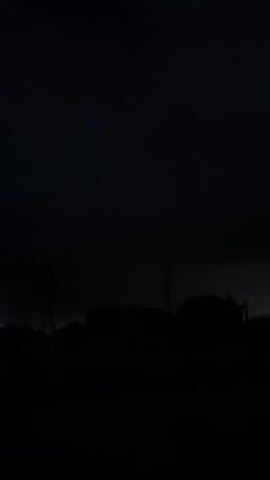 The video is a still image of a dark, silhouetted landscape. The sky is black, with faint outlines of clouds or a distant, overcast sky. Below the sky, the dark silhouettes of what appear to be buildings or structures are visible across the horizon. There are also darker, irregular shapes that could be trees or other natural elements. One tall, thin silhouette stands out on the right side of the frame, possibly a tower or a pole. The overall impression is one of nighttime or very low light conditions.