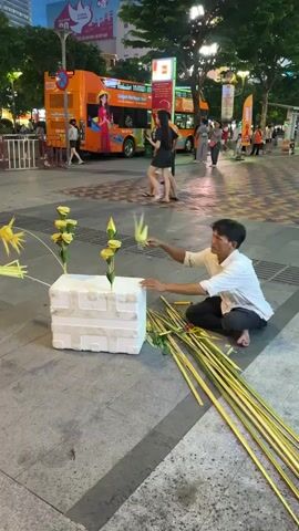 The video shows a man kneeling on a paved surface, working on an arrangement of yellow, rose-like flowers. He is using long, thin green stalks, likely palm leaves, which are bundled to his right. The flowers are inserted into a white styrofoam block. Behind him, an orange double-decker bus with advertisements is visible, along with several people walking and standing. The environment appears to be an urban outdoor setting, possibly a street or plaza, with buildings and streetlights in the background. The overall lighting suggests it is dusk or early evening.
