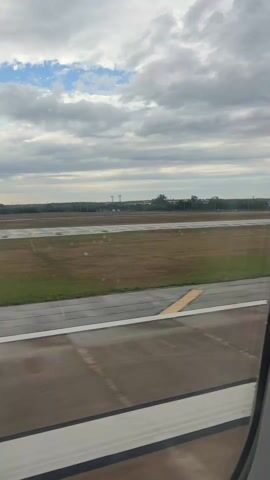 From inside an airplane, a view from the window shows a wet runway. The sky above is cloudy with patches of blue. Beyond the runway, there's a line of trees. The airplane is moving.