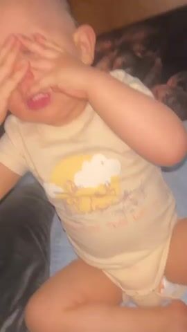 A baby wearing a light yellow onesie with a cartoon character print on the chest is shown from the chest up. The baby's hands are covering their eyes and then revealed to show a smiling expression. The background is dark and out of focus, but appears to be a bed with a patterned blanket. The lighting is dim.