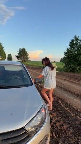 A young woman with long brown hair is seen interacting with a silver car. She is standing on a dirt road next to the open driver's side door of the car, holding a bouquet of white flowers. The background consists of a rural landscape with trees and a field under a blue sky with some clouds. The woman is wearing a light blue t-shirt and white shorts with flip-flops. She appears to be placing the flowers in the car. The video is a short clip, likely a still image with some movement.