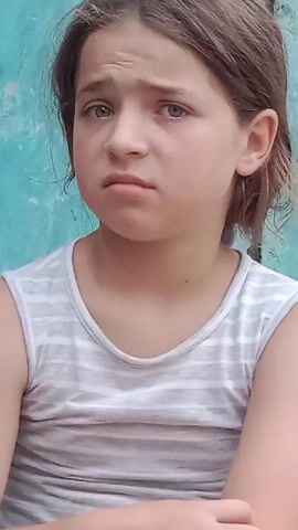 A close-up shot shows a young girl with brown hair and blue eyes looking off to the side with a concerned expression. She is wearing a gray and white striped tank top. The background is a teal-colored wall with some weathered metal fixtures. The girl's arms are crossed. The video is brief and appears to capture a moment of mild distress or apprehension.