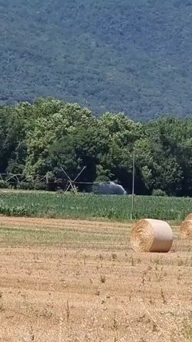 The video shows a rural landscape with fields and trees. In the foreground, there are several round hay bales. In the middle ground, there is a field of green crops and a structure that appears to be a fence or trellis. Further in the background, there is a line of trees and a large mountain covered in vegetation. There is a faint mist or spray visible in the middle ground, possibly from an irrigation system. The sky is blue and appears to be clear. The overall lighting suggests it is daytime.