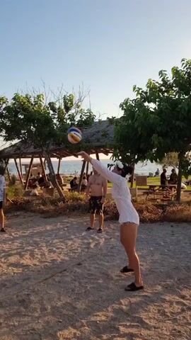 A group of four people are playing volleyball on a sandy beach. The sky is clear and blue. The sand is light brown and appears dry. The people are dressed in casual summer clothing, including shorts and t-shirts. One person in the foreground on the right is wearing a white outfit and is in the process of hitting a volleyball. Another person on the left is wearing a light-colored patterned shirt and dark shorts, and is looking up at the ball. In the background, there is a thatched-roof structure with people sitting underneath it. There are also trees and bushes surrounding the area. The ocean is visible in the distance. The volleyball is in the air, and the people appear to be engaged in a game.