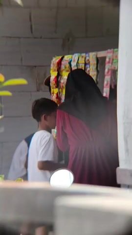 The video shows a group of people near a stall with snacks. A young boy in a white shirt is facing away from the camera, looking towards the stall. To his right, a person wearing a dark hijab and a maroon dress is also looking at the stall. To the left of the boy, another person, partially obscured, appears to be interacting with someone behind the stall, who is wearing a cap and a white garment. The stall displays rows of bagged snacks. The background consists of a light-colored brick wall. In the foreground, the reflection of a motorcycle mirror is visible. Green leaves are present in the upper left corner.