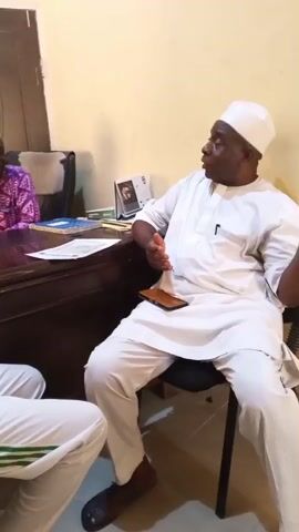 The video shows an indoor scene with several individuals in an office setting. One man, wearing a white outfit and a white cap, is seated on a chair and appears to be talking, gesturing with his hands. Another man wearing a pink patterned shirt is seated behind a wooden desk, observing the speaker. A third man, also wearing a white outfit with green stripes, is looking down at a mobile phone in his hands. A fourth person wearing a white outfit with green stripes and a green baseball cap, is looking in the direction of the speaker. The setting is a small office space, with a wooden desk, chairs, papers, and a bottle of water. A poster on the wall displays the text "Bauchi State Government of Nigeria National Anthem." The lighting is somewhat dim, and the camera pans around to show different individuals within the room.