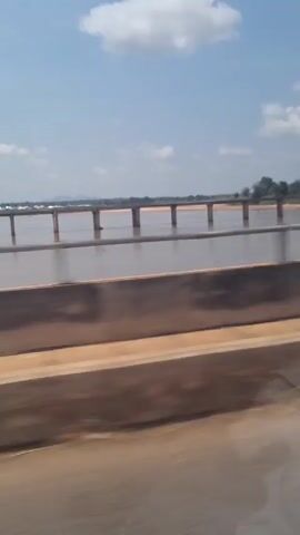 The video shows a drive across a bridge over a wide river. The camera is mounted on a vehicle that is moving.  The river is muddy, with a few exposed sandbars. The bridge appears to be a simple concrete structure with regular pillar supports. The sky is blue with scattered fluffy white clouds. The roadside has a concrete barrier. The perspective is from the passenger side of the vehicle.  The video is shaky due to the motion of the vehicle.
